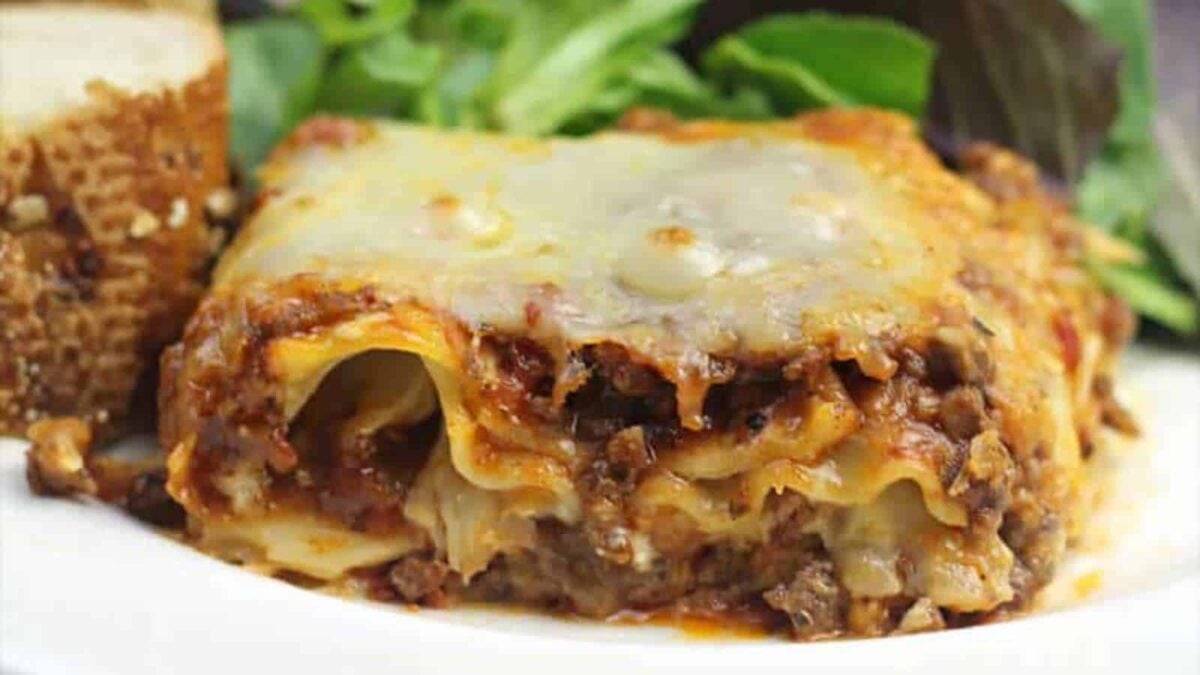 A slice of cheesy baked lasagna with visible meat layers on a white plate, served with bread and fresh green salad in the background—a must-try lasagna for every taste.