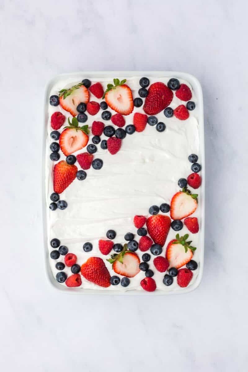 A rectangular cake topped with a layer of white frosting and decorated with strawberries, blueberries, and raspberries on a white surface.