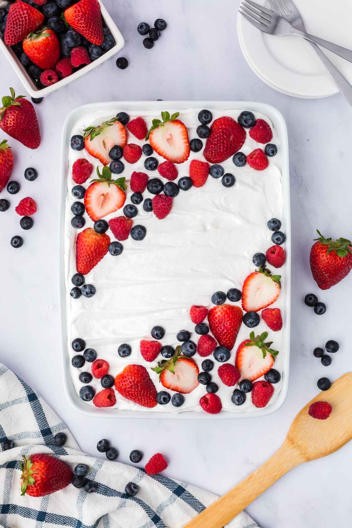Rectangular dish with a whipped cream-topped patriotic poke cake, garnished with strawberries, blueberries, and raspberries. Loose berries and serving utensils are arranged nearby on a marble surface.