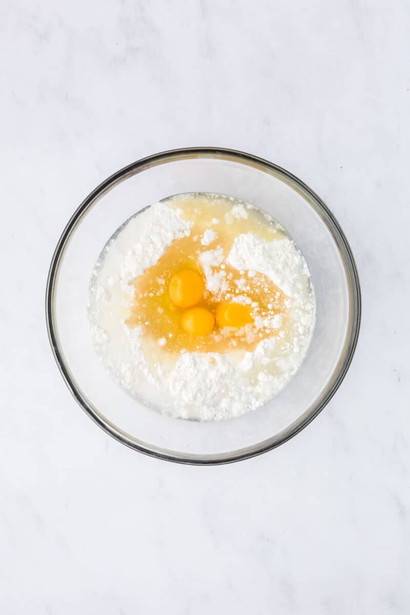 A glass bowl containing eggs, flour, and liquid ingredients on a white marble surface, ready to be mixed.