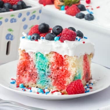 A slice of patriotic poke cake with red and blue streaks, topped with white frosting, blueberries, raspberries, and red, white, and blue sprinkles on a white plate.