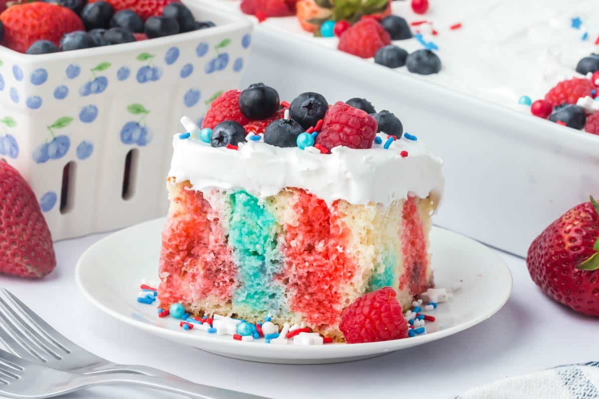 A slice of patriotic poke cake with red and blue streaks, topped with white frosting, blueberries, raspberries, and red, white, and blue sprinkles on a white plate.