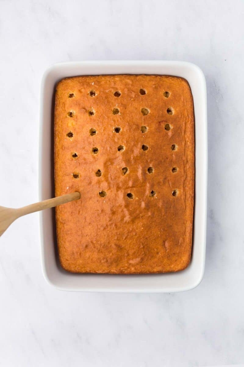 A rectangular baked cake in a white dish is being poked with holes using the end of a wooden spoon.