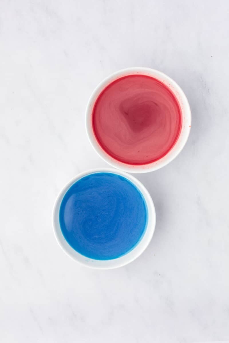 Two small white bowls, one filled with red liquid and the other with blue liquid, placed on a light marble surface.