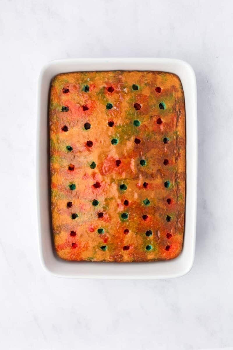 A rectangular cake in a white baking dish features evenly spaced red and green dots on top, photographed from above on a white surface.