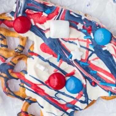 A pretzel bark coated with white, red, and blue drizzled icing, decorated with sprinkles, round candies, and mini marshmallows on a sheet of parchment paper.