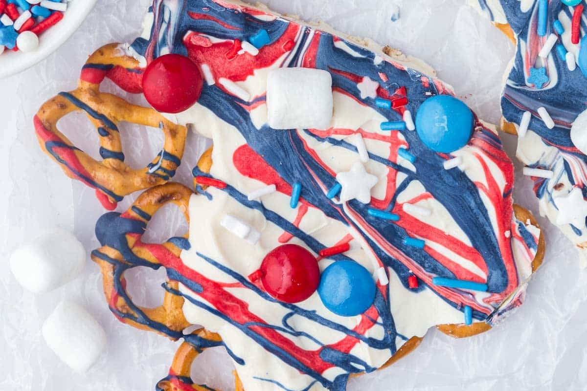 A pretzel bark coated with white, red, and blue drizzled icing, decorated with sprinkles, round candies, and mini marshmallows on a sheet of parchment paper.