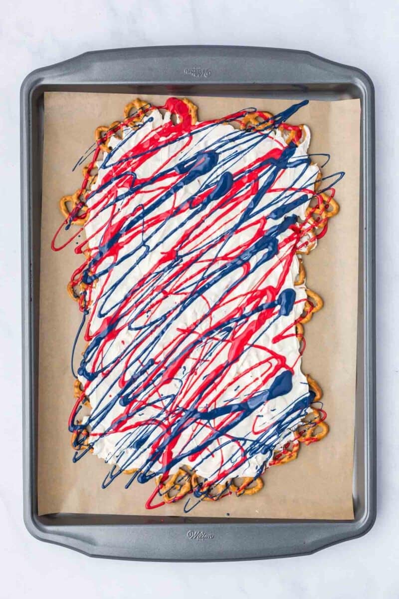 A sheet pan with a base of pretzels covered in white coating and drizzled with red and blue icing, on a piece of parchment paper.