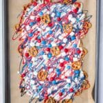 A baking tray lined with parchment holds a mix of pretzels, popcorn, red, white, and blue candies, all drizzled with red, white, and blue chocolate.