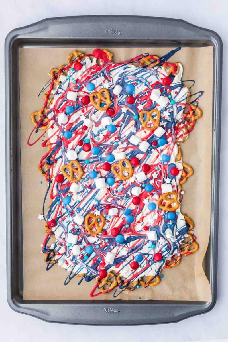 A baking tray lined with parchment holds a mix of pretzels, popcorn, red, white, and blue candies, all drizzled with red, white, and blue chocolate.