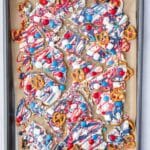 A baking sheet with pieces of white chocolate bark topped with red, white, and blue candies, pretzels, and drizzled colored chocolate.