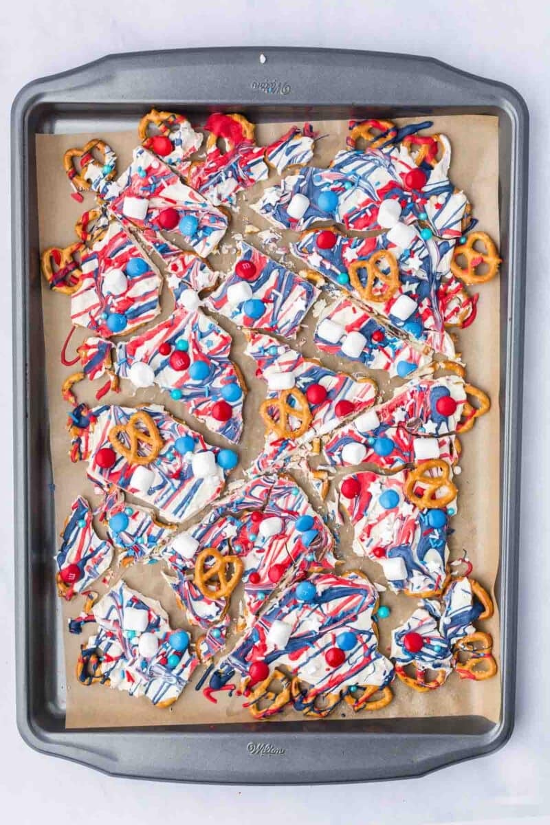 A baking sheet with pieces of white chocolate bark topped with red, white, and blue candies, pretzels, and drizzled colored chocolate.