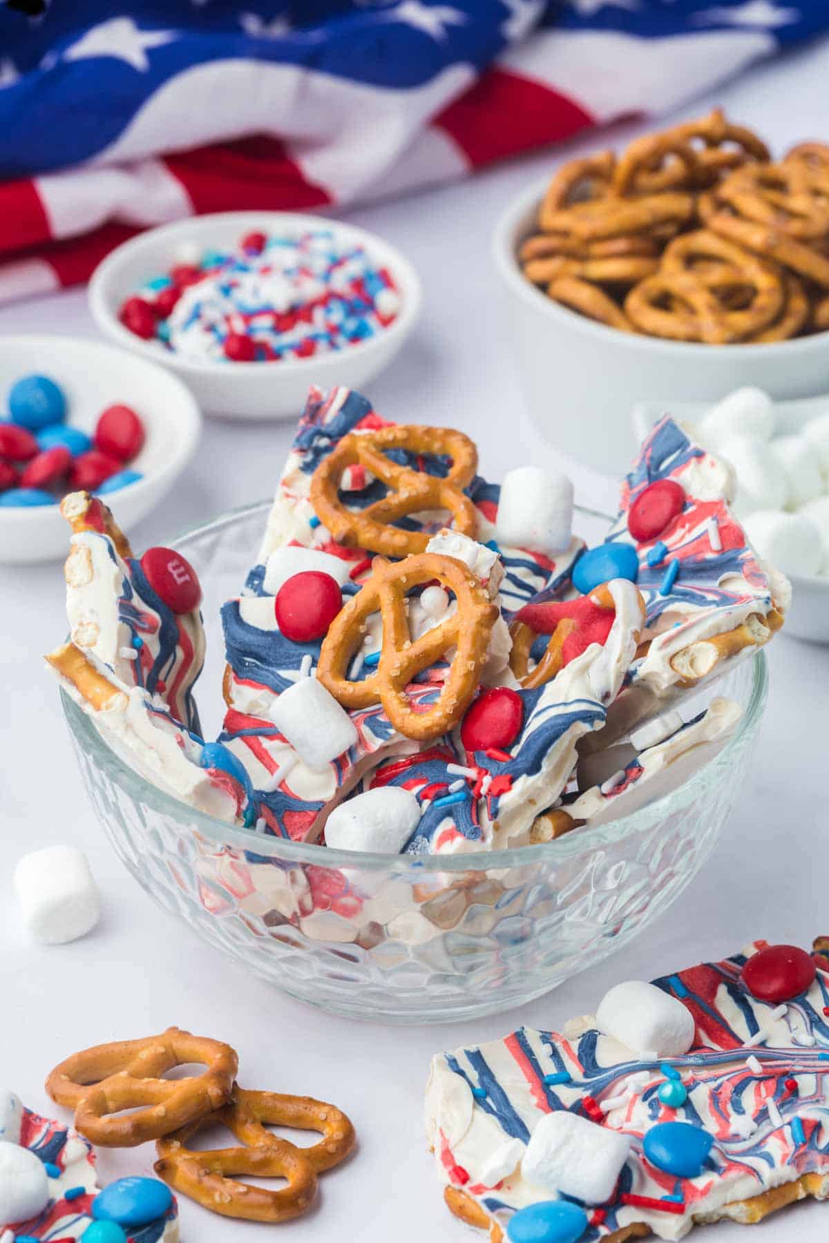 A glass bowl filled with patriotic-themed bark pieces topped with pretzels, marshmallows, and red, white, and blue candies, with a U.S. flag visible in the background.