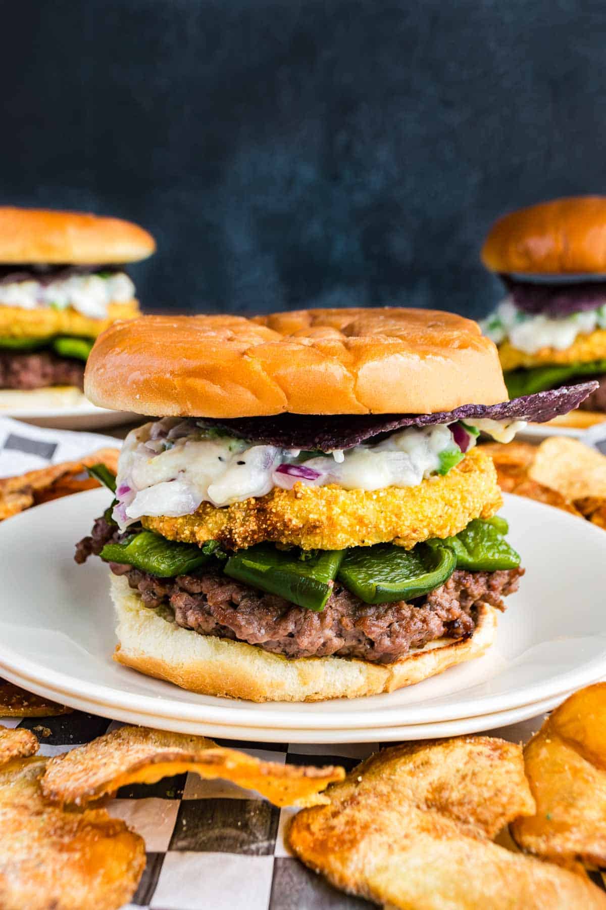 A hamburger topped with fried green tomato, leafy greens, creamy sauce, and served on a white plate with potato chips. Two similar burgers are in the background.