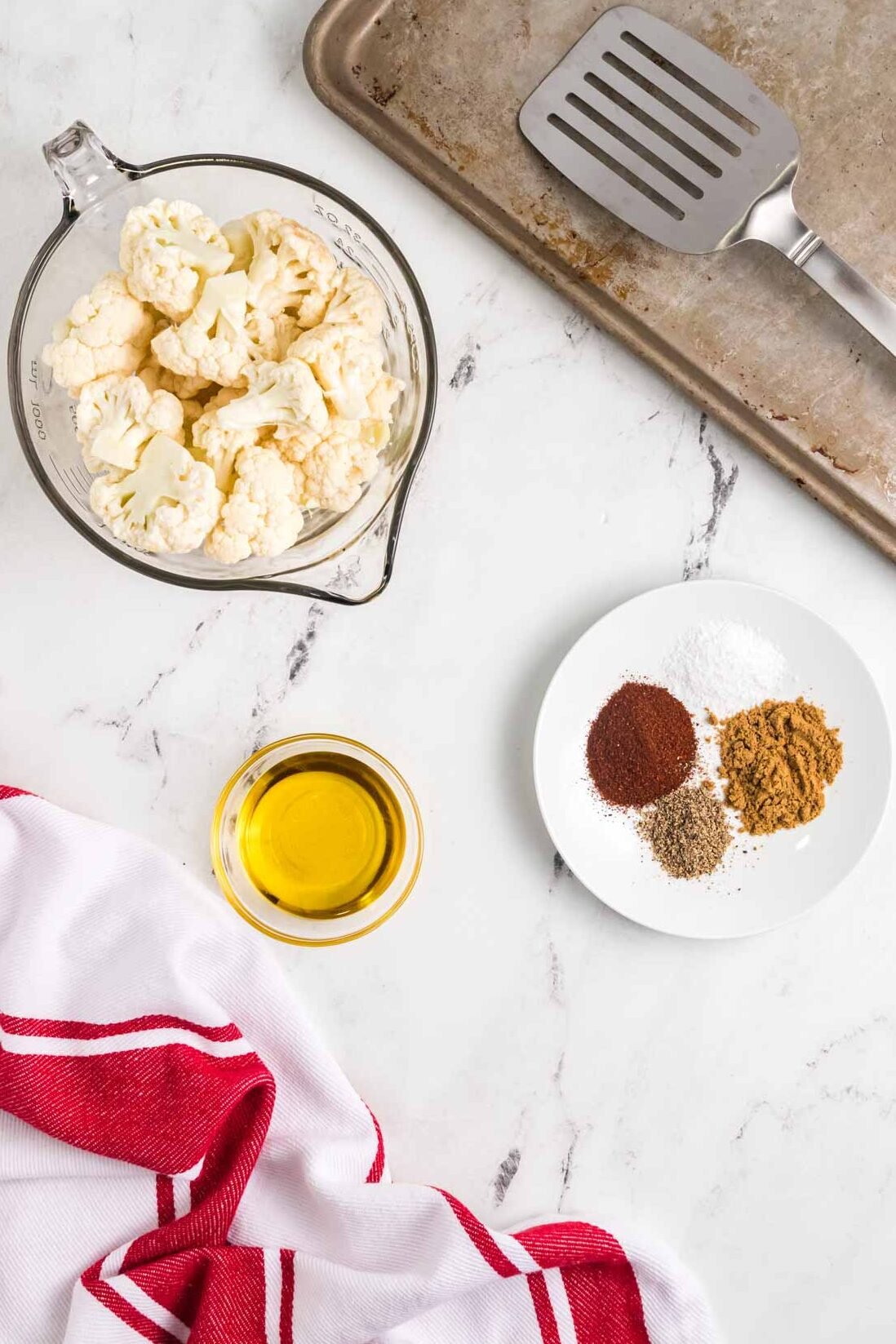 Overhead view of raw cauliflower in a glass bowl, a plate of assorted spices, a small bowl of oil, a baking sheet with a spatula, and a red and white towel on a marble surface.