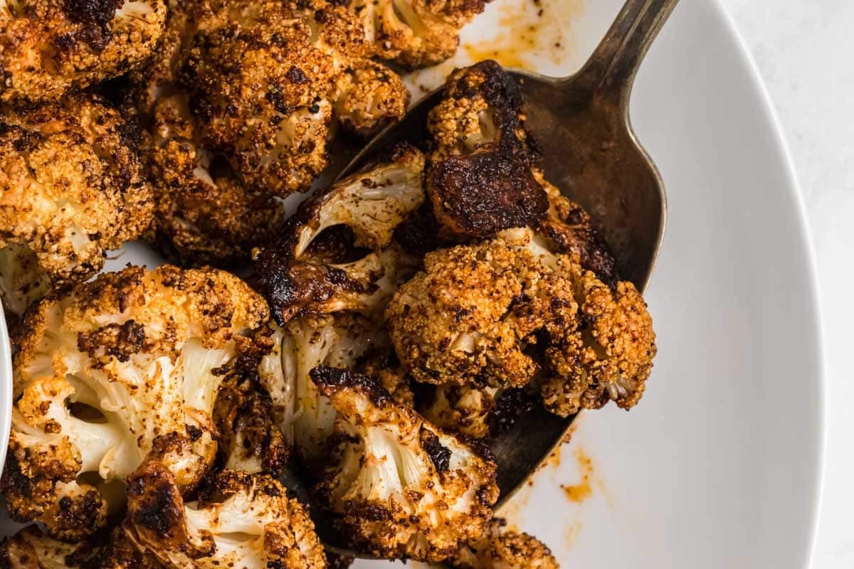 Close-up of roasted cauliflower florets with a browned, seasoned crust, served on a white plate with a metal serving utensil.