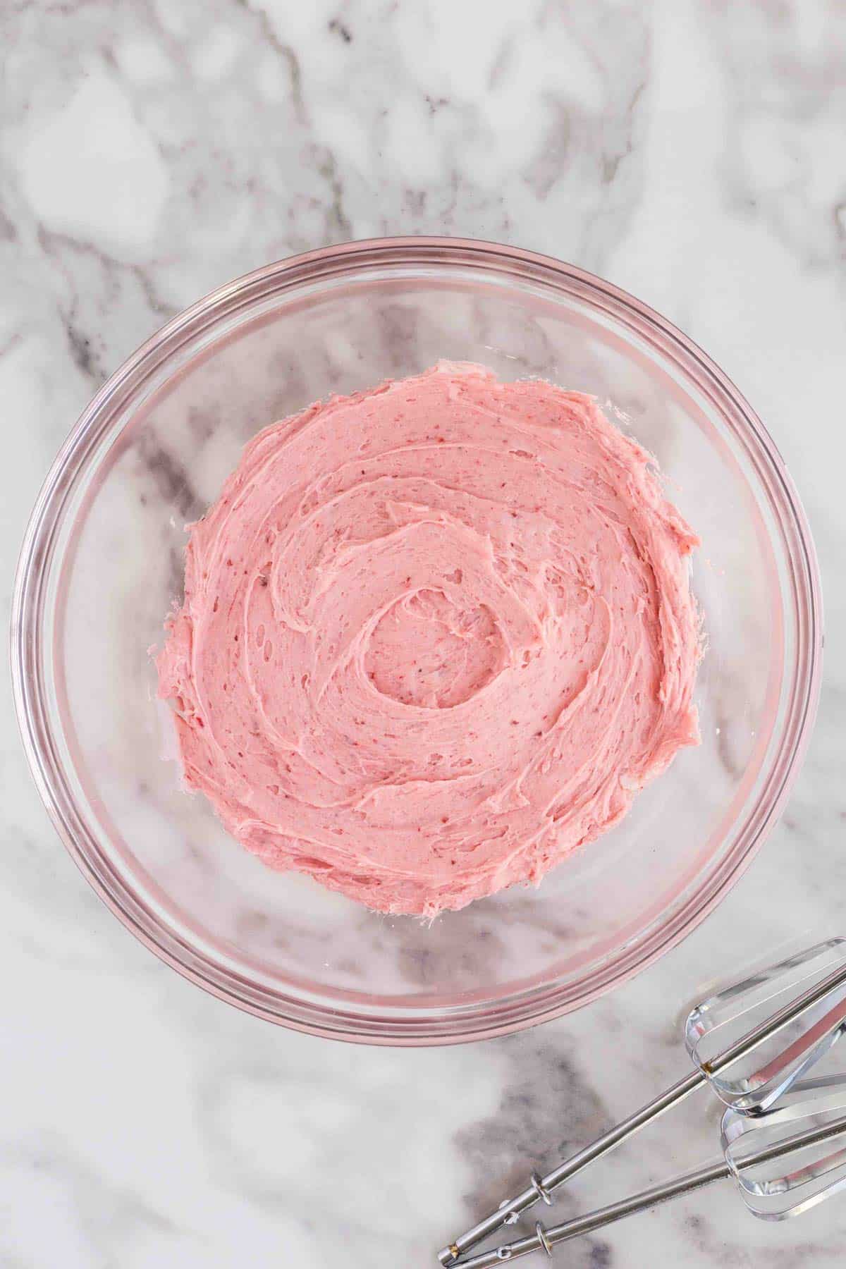 A glass bowl filled with pink whipped frosting is shown on a marble countertop. Two metal mixer beaters are placed beside the bowl.