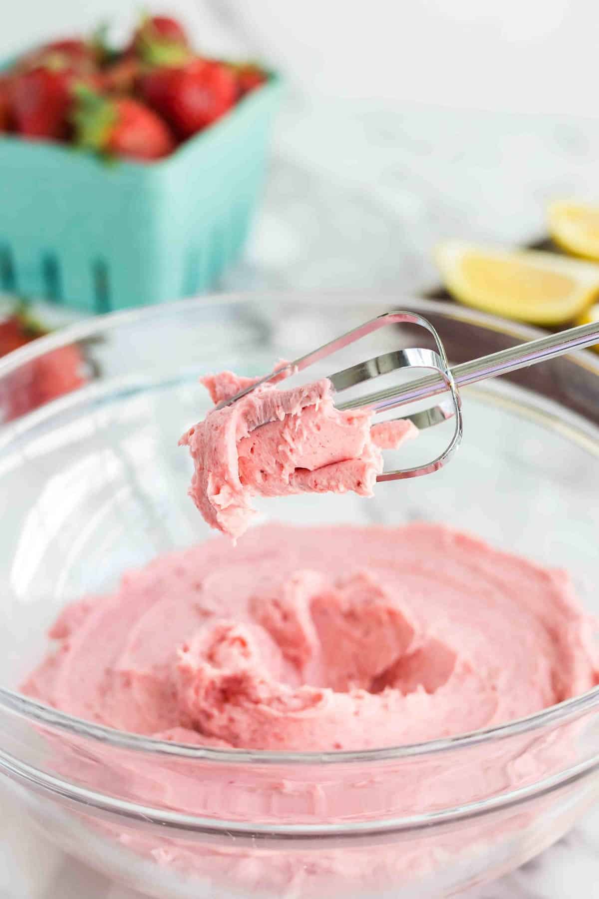 A metal beater with pink buttercream mixture is held above a glass bowl filled with more of the same mixture. A carton of strawberries and lemon slices are in the background.