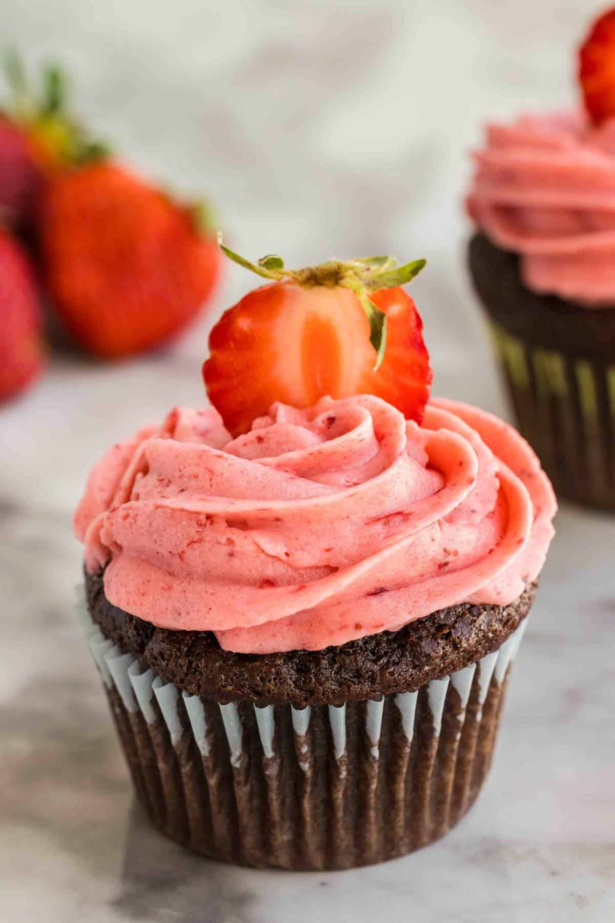 Chocolate cupcake with pink strawberry buttercream frosting, topped with a sliced strawberry, sits on a marble surface with whole strawberries in the background.