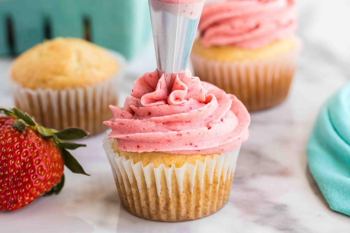 A cupcake being topped with strawberry frosting from a piping bag, with a strawberry and other cupcakes in the background.
