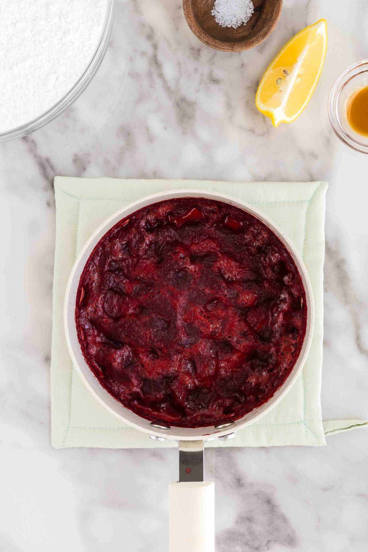 A saucepan containing thick, cooked strawberry puree sauce sits on a white mat, surrounded by a lemon wedge, a bowl of salt, and a small glass of vanilla extract on a marble surface.