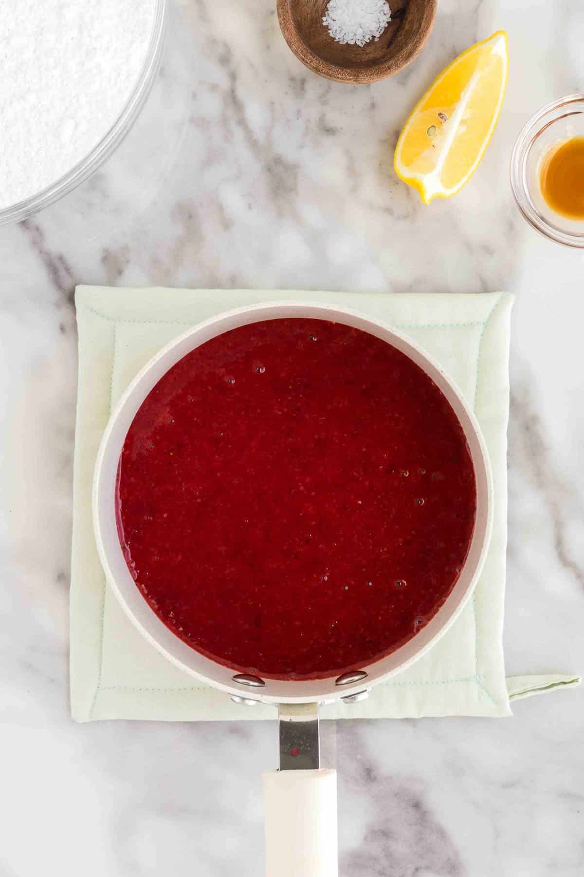 A saucepan filled with red strawberry sauce sits on a light green pad, surrounded by a lemon wedge, a small bowl of salt, and a bowl of liquid on a marble surface.