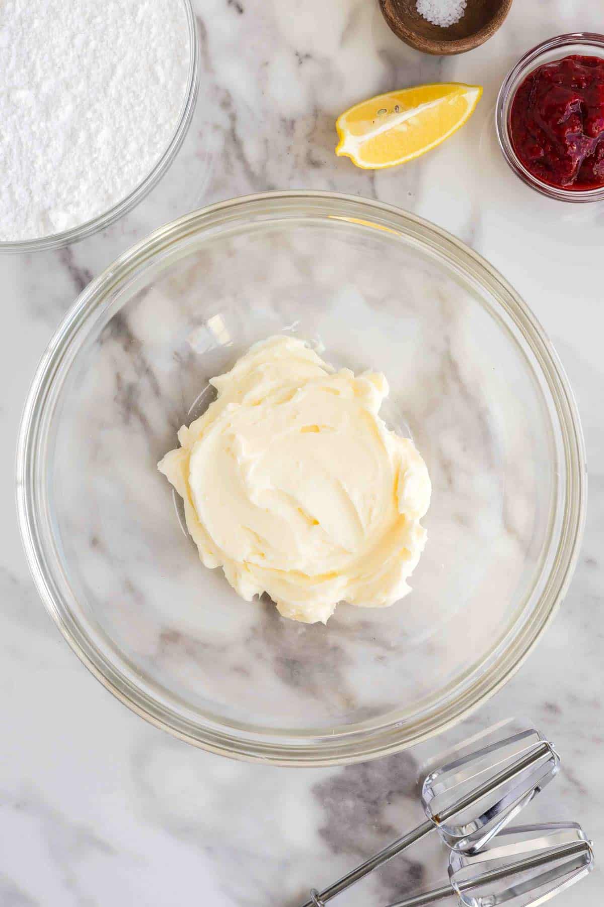 A glass bowl with creamed butter, surrounded by a bowl of powdered sugar, a lemon wedge, a small bowl of salt, a dish of jam, and a hand mixer on a marble surface.
