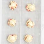 Six unbaked cookie dough portions with visible strawberry pieces and cream on top, arranged on a parchment-lined baking sheet.