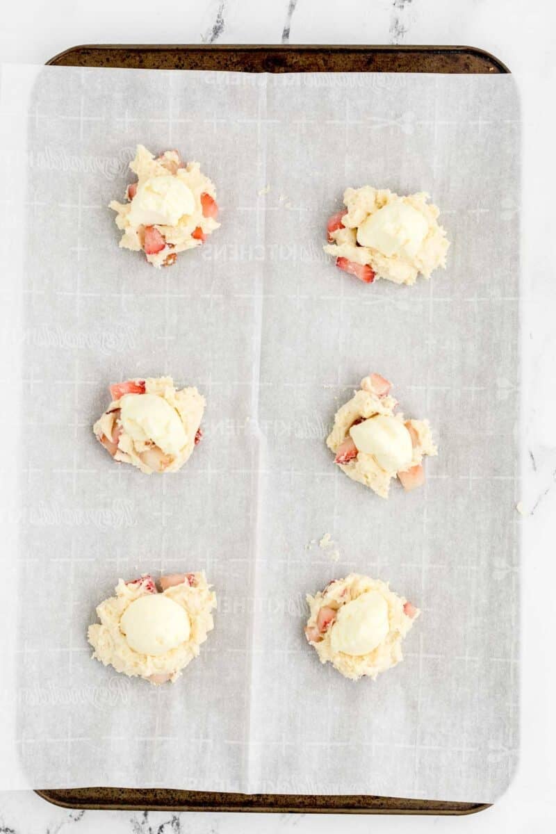 Six unbaked cookie dough portions with visible strawberry pieces and cream on top, arranged on a parchment-lined baking sheet.