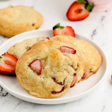 A white plate with three strawberry cheesecake cookies, garnished with fresh strawberry halves, on a marble surface.