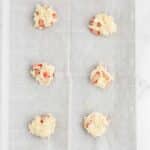 A baking sheet lined with parchment paper holds six evenly spaced scoops of raw dough with visible chunks of fruit.
