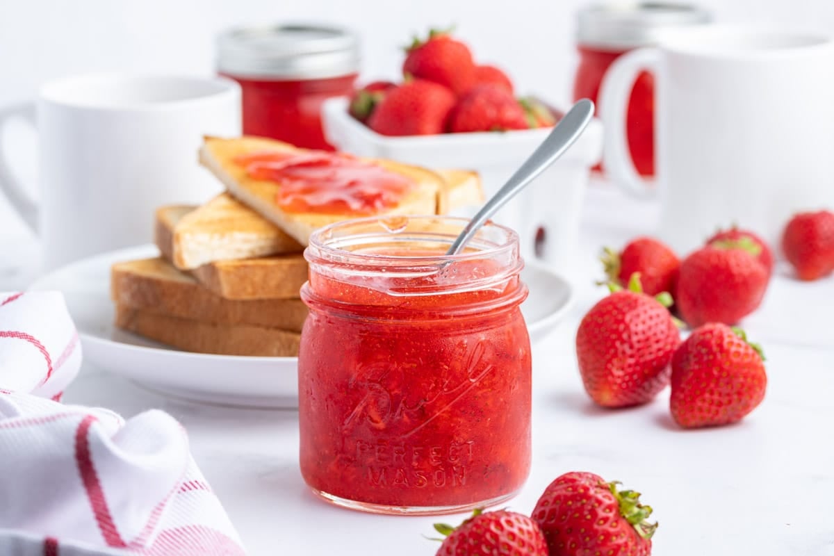 A jar of strawberry freezer jam with a spoon in it, surrounded by fresh strawberries, toast with jam, and white mugs on a white surface.