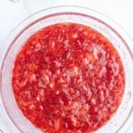A glass bowl filled with mashed strawberries on a white surface, surrounded by whole strawberries, a measuring cup of water, and a small bowl of sugar.