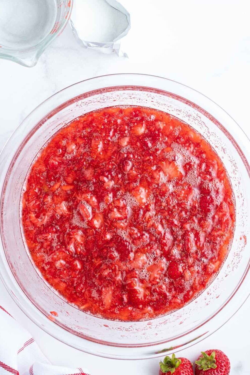 A glass bowl filled with mashed strawberries on a white surface, surrounded by whole strawberries, a measuring cup of water, and a small bowl of sugar.
