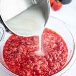 A metal pot pours a white liquid into a bowl of mashed strawberries on a white surface, with fresh strawberries and a cup of sugar in the background.