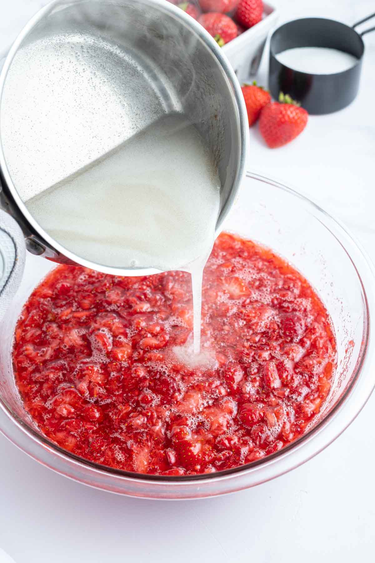 A metal pot pours a white liquid into a bowl of mashed strawberries on a white surface, with fresh strawberries and a cup of sugar in the background.