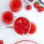 Overhead view of strawberry freezer jam being ladled into glass jars, with fresh strawberries and a bowl of sugar nearby on a white surface.