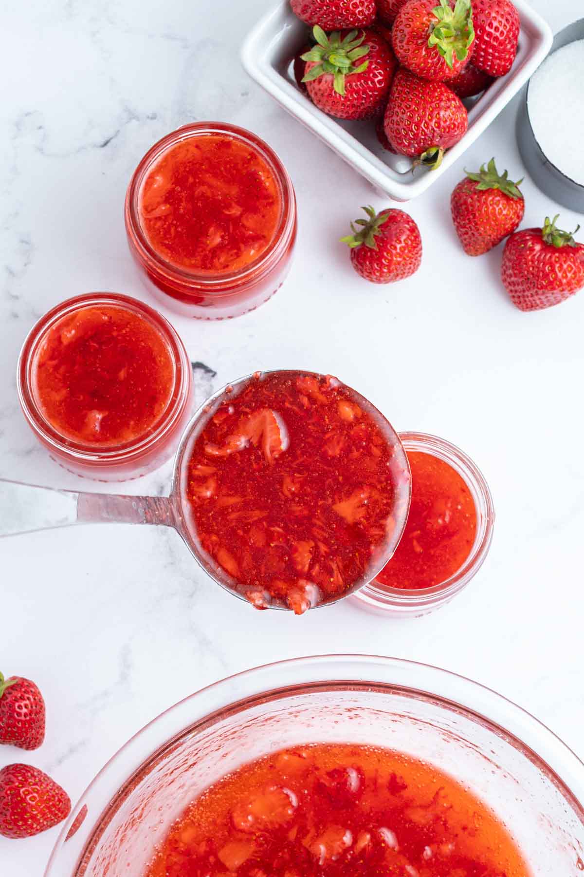 Overhead view of strawberry freezer jam being ladled into glass jars, with fresh strawberries and a bowl of sugar nearby on a white surface.