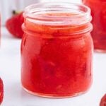 A glass jar filled with strawberry freezer jam sits on a white surface, surrounded by fresh strawberries and additional jars in the background.