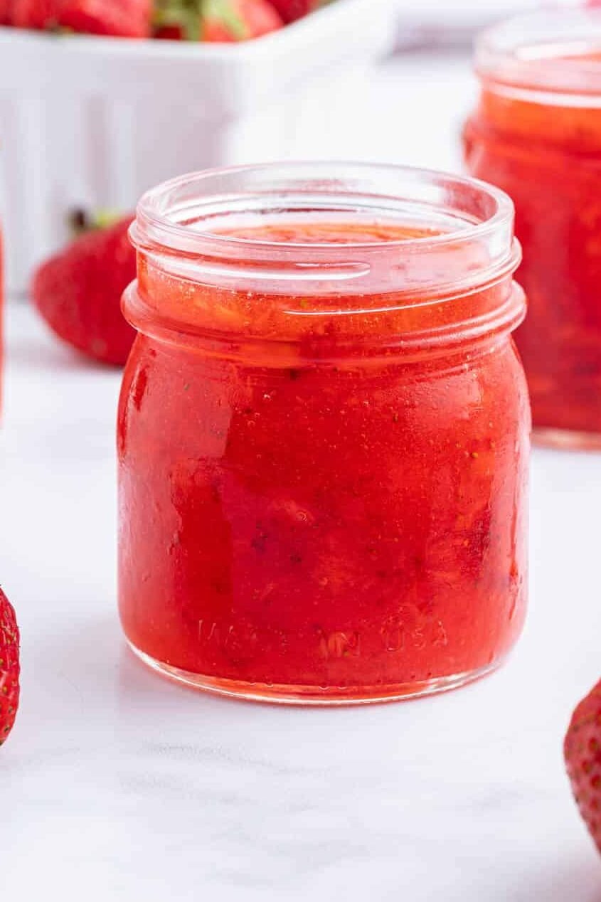 A glass jar filled with strawberry freezer jam sits on a white surface, surrounded by fresh strawberries and additional jars in the background.