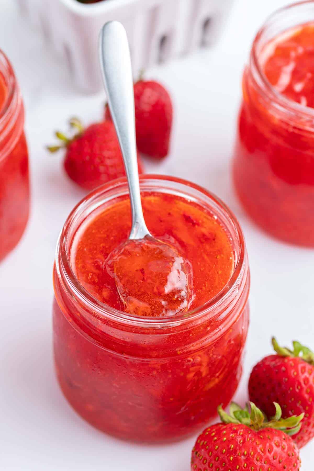 A glass jar filled with strawberry freezer jam, with a spoon resting inside, surrounded by fresh strawberries on a white surface.