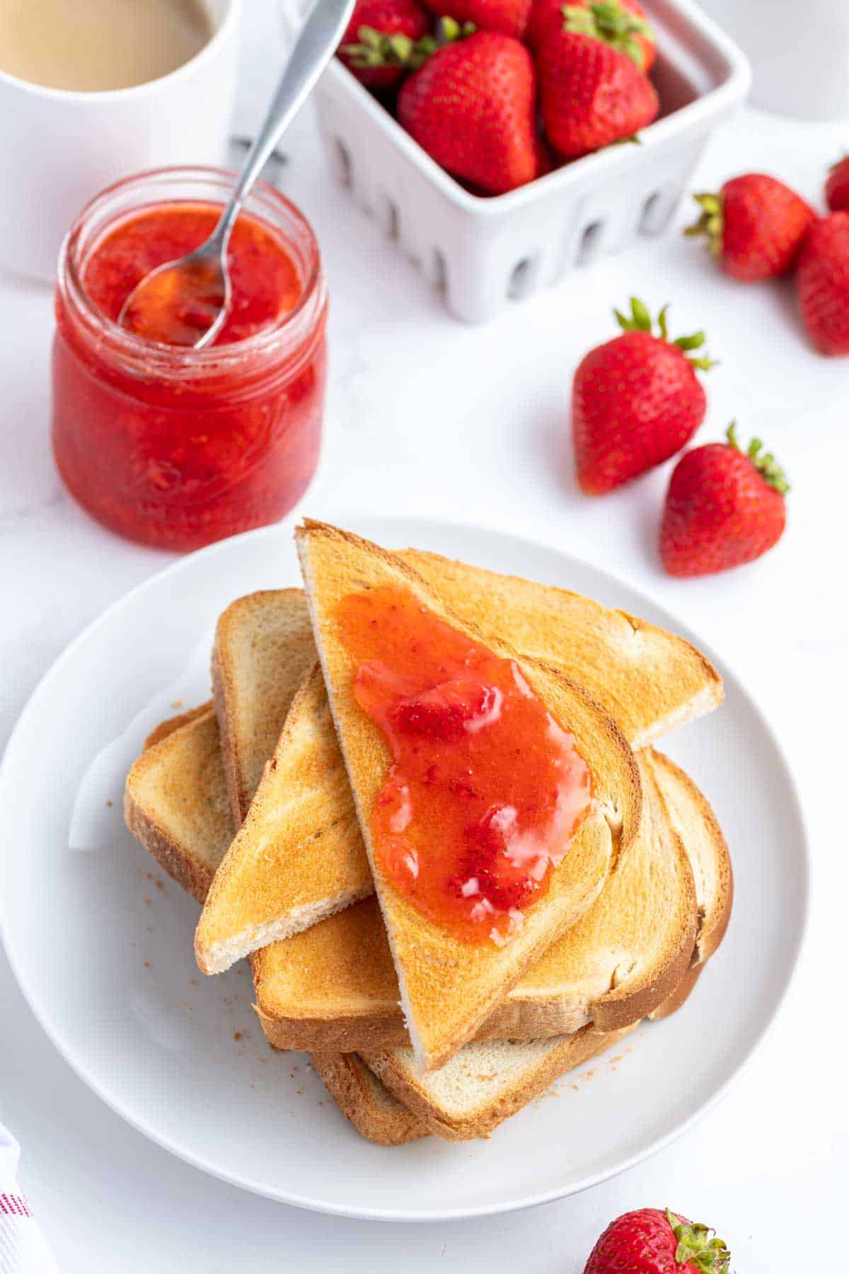 A plate of toasted bread slices with one slice topped with strawberry freezer jam, surrounded by fresh strawberries, a jar of jam, and a cup of coffee.