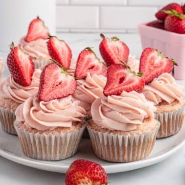 A plate of cupcakes with strawberry frosting with jam and halved strawberries on top, with whole strawberries and a basket of strawberries in the background.