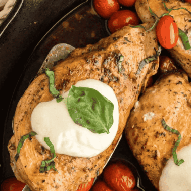 Two seasoned chicken breasts in a skillet topped with melted mozzarella, fresh basil, and surrounded by cherry tomatoes.