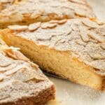 A close-up of a sliced almond cake topped with powdered sugar and almond flakes.