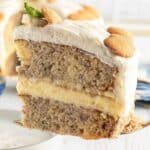 A slice of banana pudding cake with creamy filling, frosted with white icing, and topped with vanilla wafer cookies and a mint leaf.