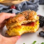 A hand holds a hawaiian roll breakfast slider with egg, bacon, and cheese on a seeded bun, showing a bite taken out. Berries are on the plate nearby.