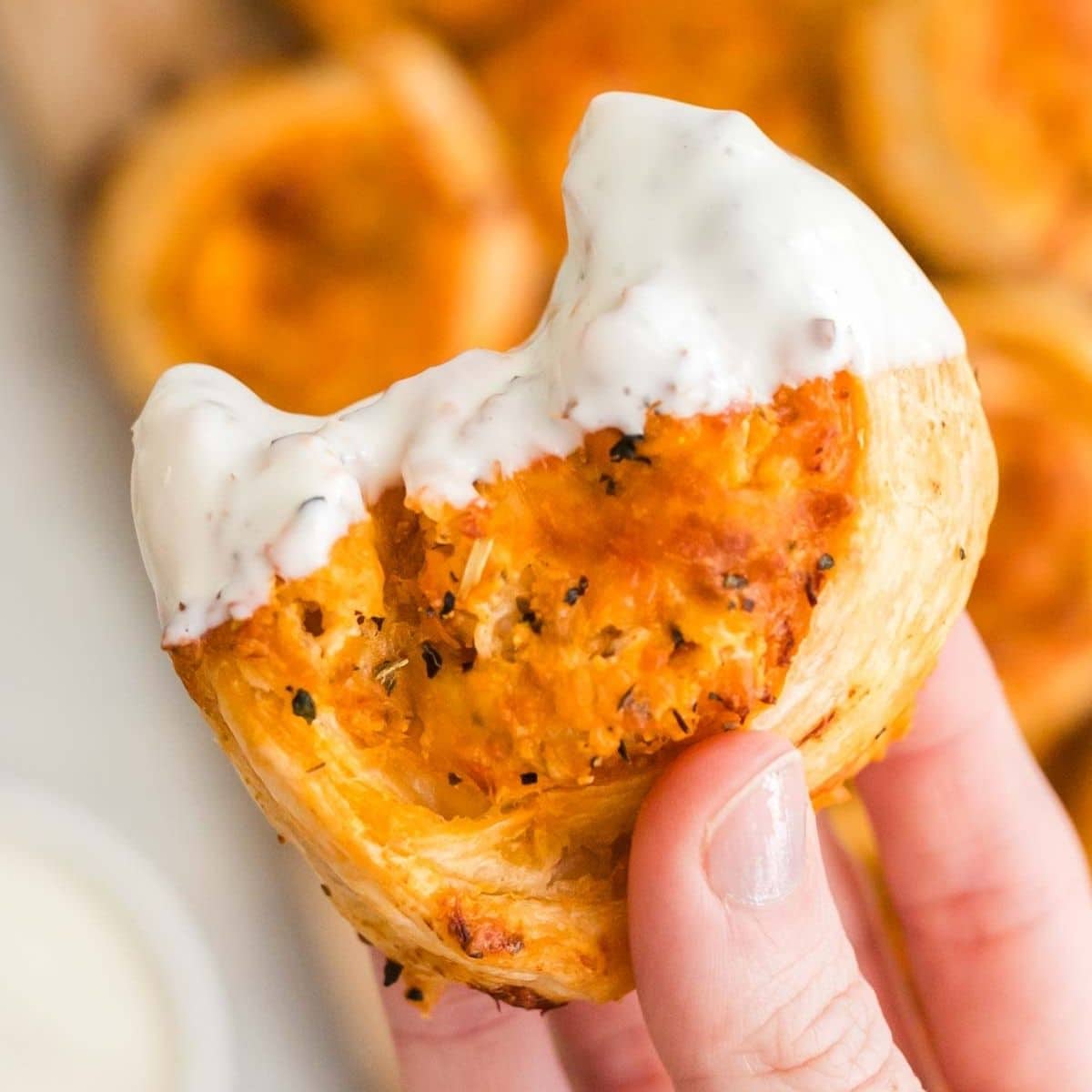 A hand holds a partially eaten, saucy, bite-sized buffalo chicken pinwheel with a flaky crust and creamy white dip on top.