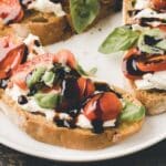 Toasted bread topped with ricotta cheese, cherry tomatoes, fresh basil leaves, and a drizzle of balsamic glaze on a white plate.