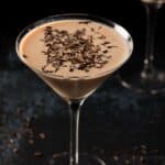 A martini glass filled with a chocolate martini with baileys cocktail, topped with chocolate shavings, set against a dark background.